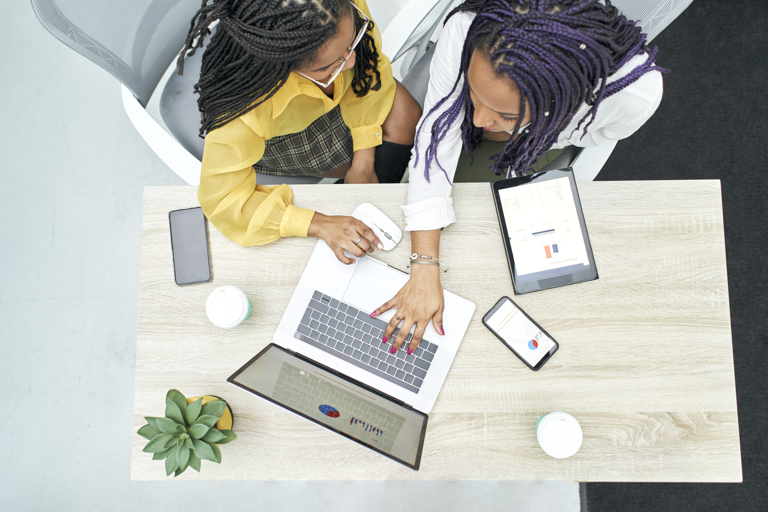 Top view of two young black women working in the office. Reviewing analytical data on various electronic devices. Zenith view.