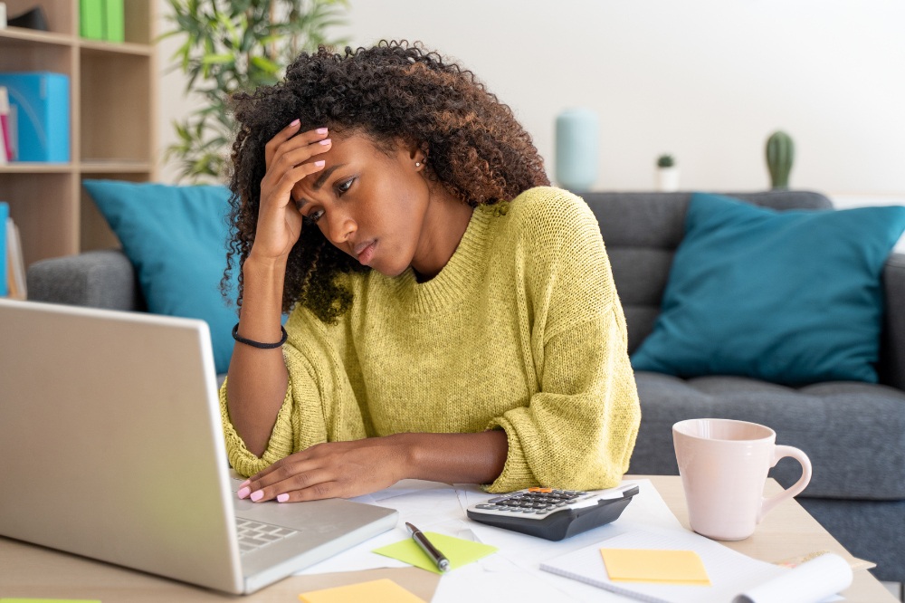 A black woman frustrated by scope creep, is reviewing a project scope document on her laptop, with papers and coffee cup on the side.